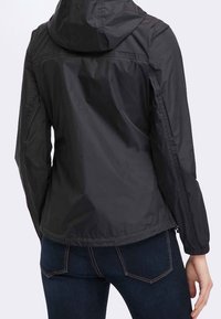 Donna che indossa una leggera giacca nera con cappuccio e jeans blu scuro, vista da dietro su uno sfondo neutro.