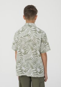 Camicia a maniche corte con bottoni in tessuto verde con un motivo di foglie tropicali bianche. Collo dritto e vestibilità rilassata. Mostrata la vista posteriore.