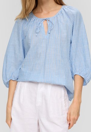 Woman wearing a blue and white vertical striped blouse with puffed sleeves and tie detail at the neckline, paired with white pants.