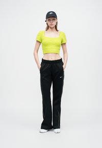 Pulover scurt verde lime cu mâneci bufante, asortat cu pantaloni de trening negri cu dungi albe pe laterale și o şapcă neagră Nike.