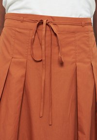 Rust-colored skirt with pleats and a tied fabric belt at the waist, worn with a white top.