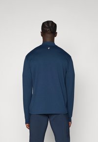 Navyblaue Langarmshirt aus weichem Stoff, mit Rundhalsausschnitt und einem kleinen Logodetail auf der Rückseite. Getragen mit passenden Hosen.