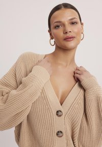 Beżowy, oversize kardigan z dekoltem w kształcie litery V, teksturowanym dzianinowym materiałem, dwoma dużymi guzikami i ściągaczami na mankietach. Gładka, miękka tekstura z luźnym krojem.