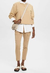 Beige sweatshirt med präglad "ESPRIT"-logga, lagd över en vit skjorta. Beige cargobyxor med fickor och svarta sandaler med vita remmar.