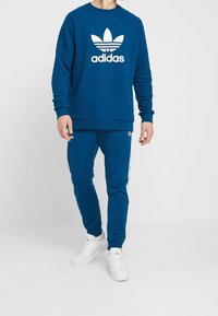 Sweatshirt bleu avec logo Adidas blanc et trois bandes, associé à un pantalon de jogging bleu assorti. Des baskets blanches complètent la tenue.