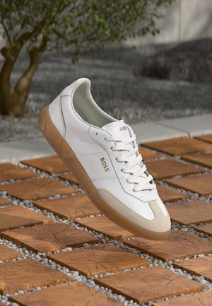 Białe skórzane trampki z beżowymi zamszowymi akcentami, z okrągłym czubkiem, sznurowanym przodem i brązową gumową podeszwą, z widoczną nazwą marki "BOSS."