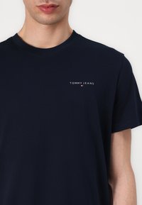 T-shirt en coton bleu marine avec un col rond, arborant un petit logo blanc "TOMMY JEANS" sur la poitrine gauche et une texture lisse.