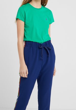 Jeune femme portant un t-shirt vert à manches courtes et un pantalon bleu marine à cordon avec des bandes latérales multicolores, debout devant un fond uni.