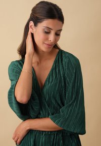 Grünes plissiertes Kleid mit tiefem V-Ausschnitt und weiten Ärmeln. Der Stoff hat eine strukturierte Oberfläche, die eine fließende Silhouette erzeugt. Einfaches goldenes Armband als Akzent.