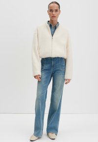 Veste zippée en crème texturée avec des manches larges, associée à un jean bleu à taille haute et jambes larges, et des chaussures plates beiges.
