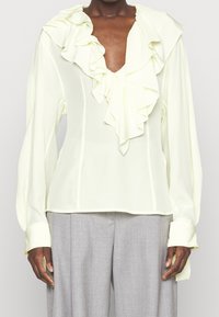 Person som bär en blekgul blus med volanger och långa ärmar, samt vida grå byxor mot en enfärgad bakgrund.