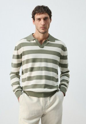 Hombre con las manos en los bolsillos, lleva una camiseta polo de manga larga a rayas verdes y blancas y pantalones de color claro, sobre un fondo liso.