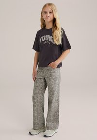 Juodas marškinėliai su užrašu "JAUNAS IR LAISVAS", derinami su plačiomis leopardų rašto kelnėmis. Visumą užbaigia šviesios spalvos sportbačiai.