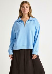 Lichtblauwe zip-up sweater met een brede kraag, verlaagde schouders en geribde manchetten, gedragen over een zwarte vloeiende rok. Gladde textuur.