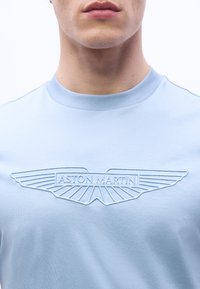 Κοντινό πλάνο ενός ατόμου που φοράει ένα γαλάζιο πουκάμισο με ανάγλυφο λογότυ πο Aston Martin στο στήθος, εμφανίζοντας μόνο τα χείλη και τον λαιμό.