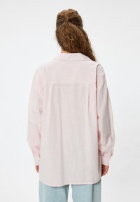 Lyserød oversized skjorte med lange ærmer og en knaplukning ved manchetten. Har et tekstureret stof og en krave, samt en let buet kant.