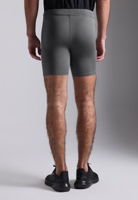 Grå treningshorts med en glatt tekstur, tettsittende design og elastisk midje, sett fra baksiden. Svart sko er brukt.