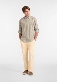 Camicia a righe beige e marrone, pantaloni color crema, scarpe marroni senza lacci e un piccolo dettaglio con logo sulla camicia.