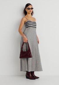 Γυναίκα με μαξι μεγάλο φόρεμα σε σχέδιο ασπρόμαυρο, σκούρα γυαλιά ηλίου, καφέ μπότες και κρατώντας καφέ τσάντα που ταιριάζει, στέκεται μπροστά σε λευκό τοίχο.