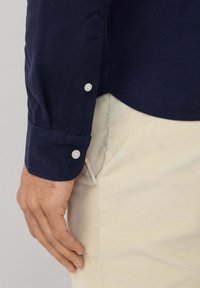 De linkerhand van de man rust nabij de voorzak, terwijl hij een marineblauw overhemd met lange mouwen en witte knopen draagt, met beige broeken.