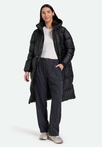 Cappotto lungo imbottito nero con colletto alto, tasche con zip e polsini elasticizzati, indossato sopra una maglietta bianca e pantaloni a righe verticali. Sneakers grigie chiare.