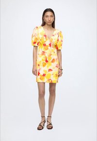 Femme portant une robe courte jaune à fleurs avec des manches bouffantes et des talons noirs à brides, debout devant un fond blanc uni.
