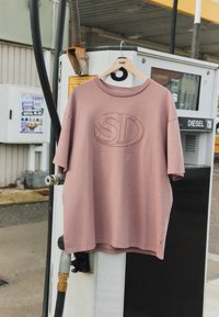 T-shirt rose poussiéreux avec logo "SD" sur un cintre en bois, suspendu à une pompe à essence diesel dans une station-service.