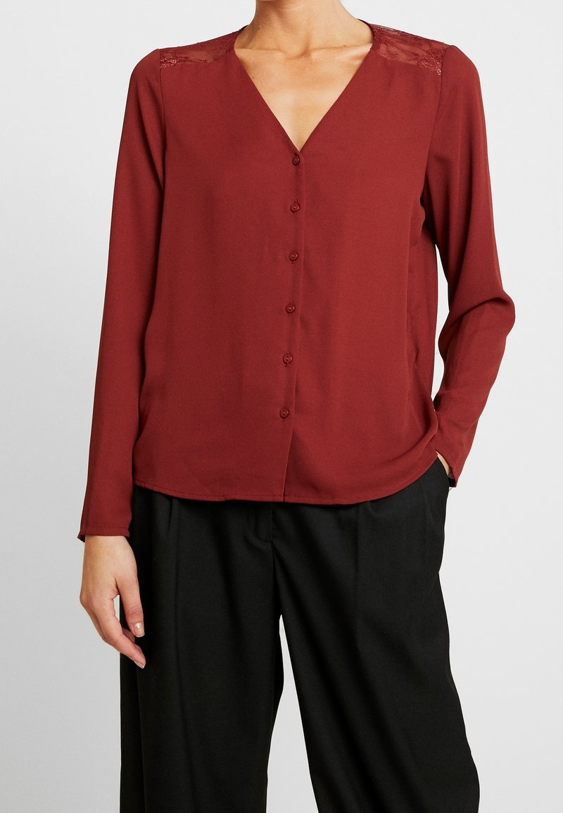 Blouse boutonnée bordeaux à manches longues, col en V, avec détail en dentelle sur les épaules, fabriquée en tissu lisse, associée à un pantalon noir.