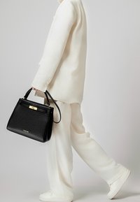 Borsa a mano nera in pelle con una forma strutturata, accessori in oro e manico superiore, tenuta da una persona in un completo bianco e sneakers.