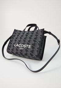 Czarna torba o teksturowanej powierzchni z geometrycznym wzorem, z białym logo "LACOSTE 1933" oraz regulowanym czarnym skórzanym paskiem i uchwytami.