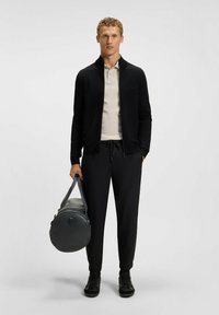 Schwarze Reißverschlussjacke, beiges Polohemd und schwarze Jogginghosen. Hält eine runde graue Sporttasche. Strukturiertem Material auf der Jacke, elegantes Design. Schwarze Sneaker.