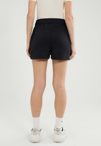 Persona de pie de espaldas, vistiendo pantalones cortos elásticos negros, calcetines blancos y zapatillas deportivas blancas contra un fondo liso.