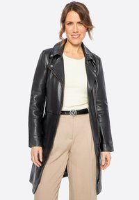 Abrigo de cuero negro con un diseño ajustado, cremallera asimétrica y solapa clásica. Llevado sobre una blusa de color claro y pantalones beige.