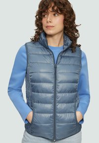 Blauer Puffy-Weste mit hohem Kragen, gestepptem Design und Reißverschluss. Getragen über einem hellblauen Langarmshirt. Texturierte, glatte Oberfläche.