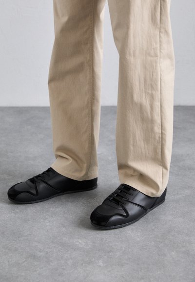 Black sneakers feature a smooth leather and suede upper, flat laces, and a textured rubber sole. Paired with beige pants against a gray floor.