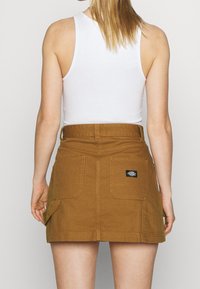 Jupe en denim marron avec une taille ajustée, dotée de deux grandes poches arrière et d'une étiquette logo noire. Le design est décontracté et utilitaire.