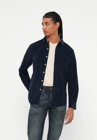 Camisa de botones navy de tela de pana, con un bolsillo en el pecho y textura suave, combinada con una camiseta beige clara y jeans oscuros.