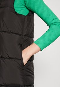 Schwarze Steppweste mit quiltetem Design und einer Seitenasche, getragen über einem grünen langärmligen, gerippten Oberteil. Glatte Textur, enganliegende Silhouette.