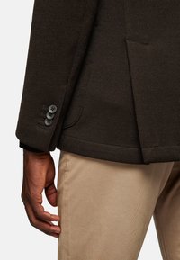 Dark brown textured jacket with three-button cuff detail, paired with light beige trousers. The jacket has a smooth finish and structured shape.