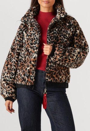 Femme portant une veste en fausse fourrure imprimée léopard, un pull rouge et un jean foncé, tenant le tirette de la fermeture éclair de la veste avec une étiquette rouge.