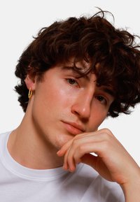 Boucle d'oreille en or mise en valeur sur un mannequin aux cheveux bouclés et foncés, portant un t-shirt blanc à col rond ; gros plan sur les traits du visage.
