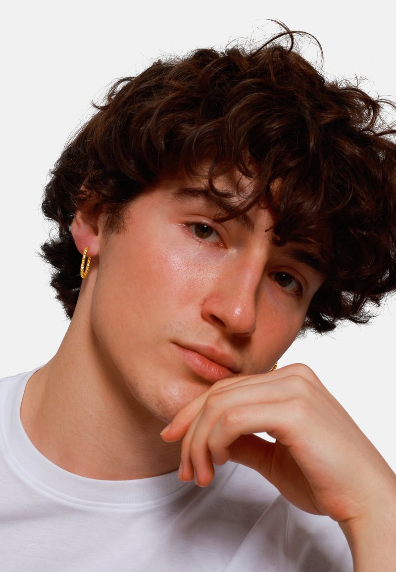 Boucle d'oreille en or mise en valeur sur un mannequin aux cheveux bouclés et foncés, portant un t-shirt blanc à col rond ; gros plan sur les traits du visage.