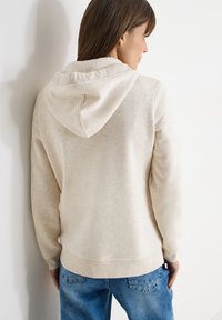 Femme aux cheveux bruns portant un sweat à capuche beige clair et un jean bleu, vue de dos contre un mur blanc.