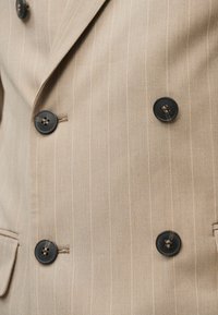 Μπεζ διπλόμπουτο σακάκι με ρίγες, με τέσσερα μαύρα κουμπιά και μια πλαϊνή τσέπη ορατή στο κάτω αριστερό μέρος.