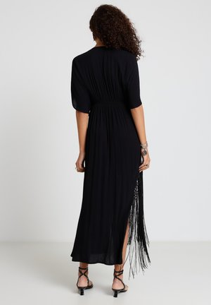 Donna con capelli ricci che indossa un vestito nero lungo fino alla caviglia con maniche corte, spacco laterale, dettagli frangiati, tacchi neri con cinturini e braccialetti argentati.