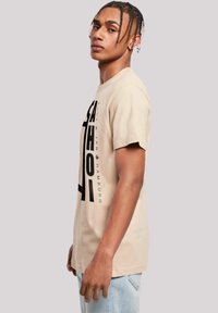 Beige Baumwoll-T-Shirt mit kurzen Ärmeln, das auf der Vorderseite mit fettem schwarzen Grafiktext versehen ist. Es hat eine lockere Passform und einen Rundhalsausschnitt.