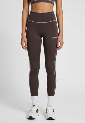 Kvinde iført brune højtaljede leggings med hvide syninger, hvide sokker og hvide sneakers, stående mod en ensfarvet baggrund.