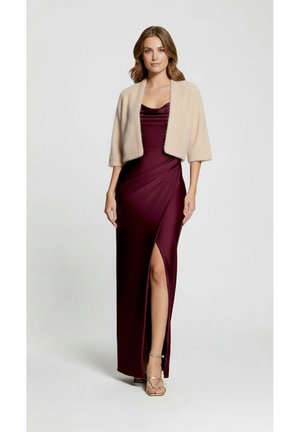 Femme portant une robe de soirée en satin bordeaux avec une fente à la cuisse et un cardigan court beige, debout devant un fond blanc uni.