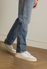 Weiße und beige Sneakers mit einem strukturierten Design und Schnürung, kombiniert mit hellblauen Jeans, auf einem Holzboden.