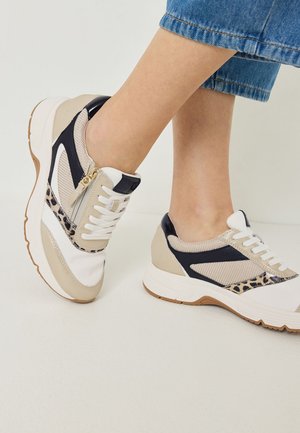 Füße in beigen, weißen und schwarzen Sneakers mit Leopardenmuster-Akzenten und einem Reißverschlussdetail, kombiniert mit cropped blauen Jeans.